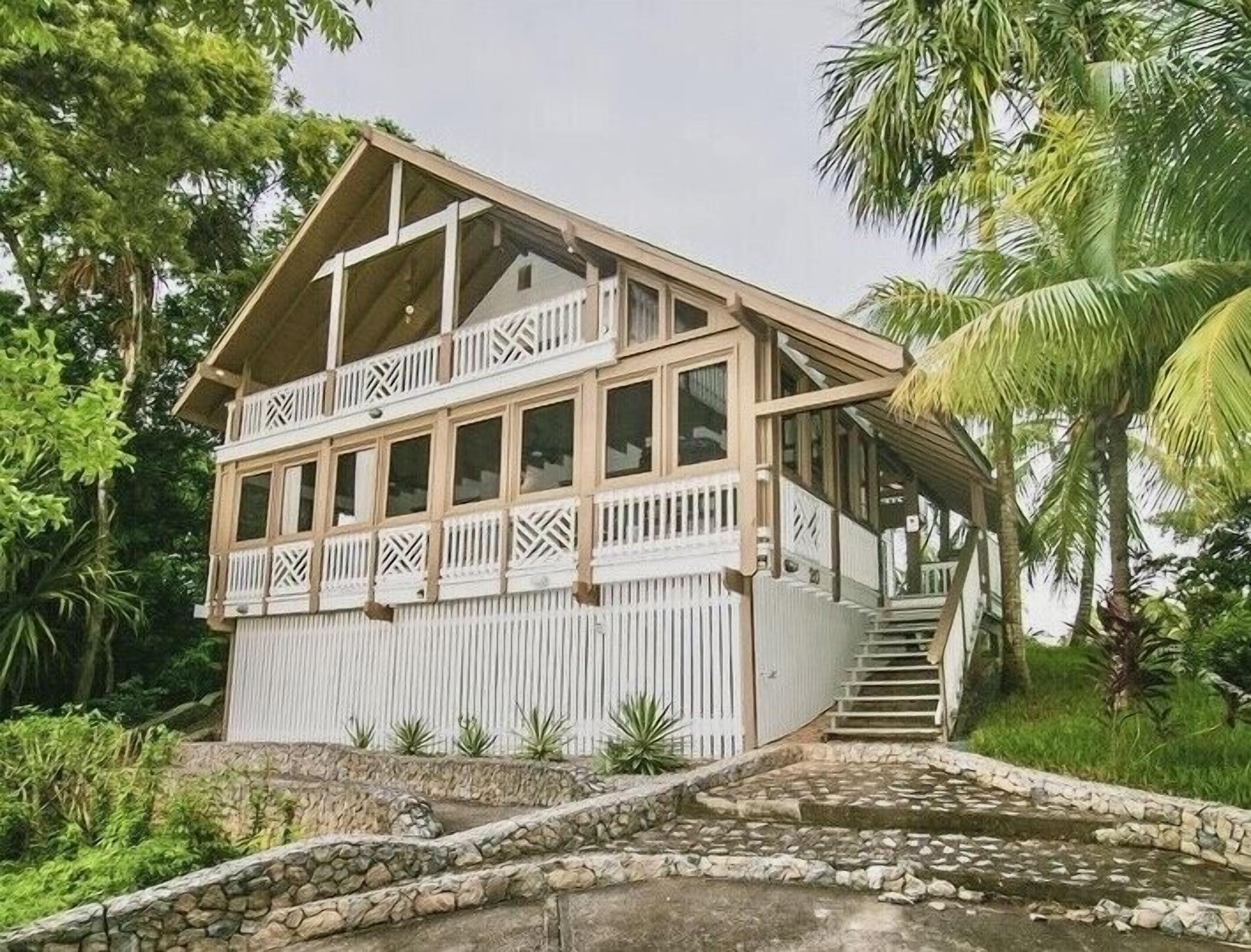 A two-story wooden house with a balcony, surrounded by tropical vegetation.