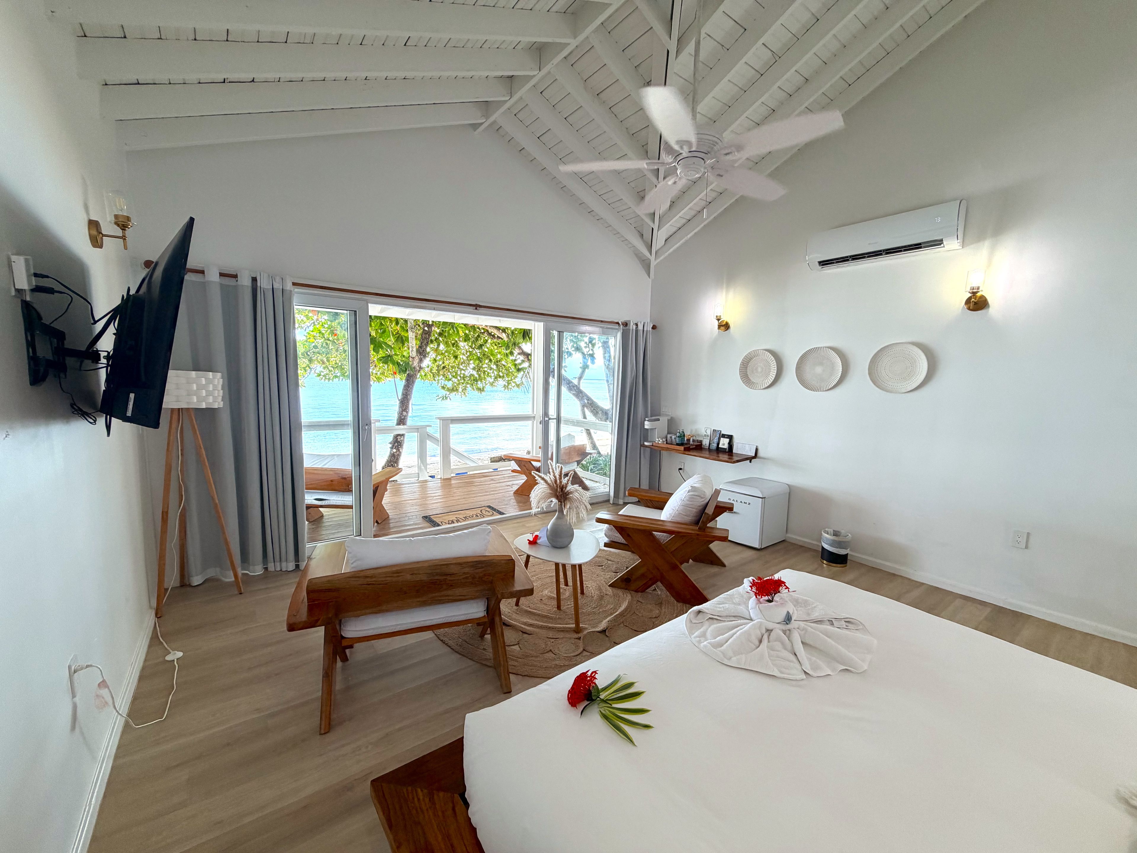 A room with a bed, a desk, a chair, a ceiling fan, and a view of the sea through a sliding door.
