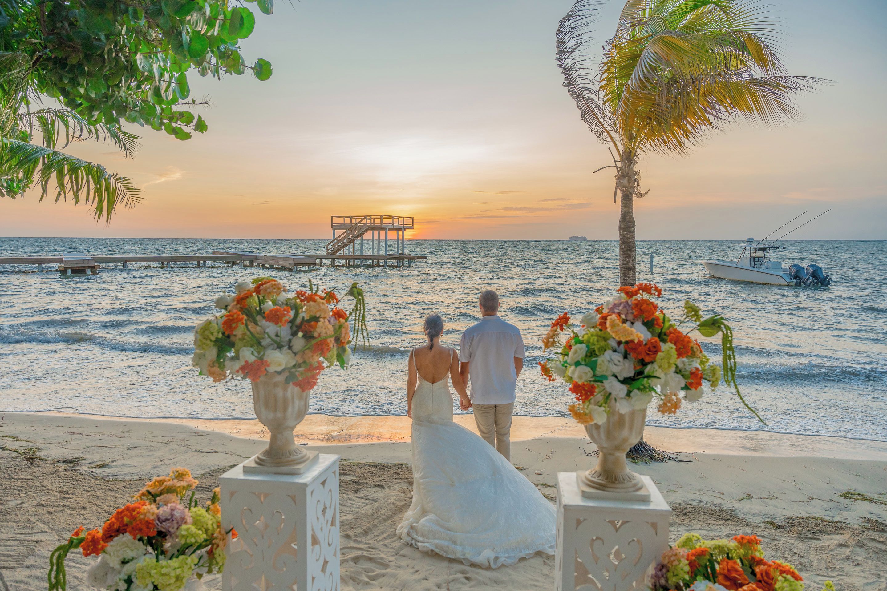 Ceremonia de boda en la playa con una pareja tomándose de las manos, un muelle al fondo y un barco en el agua.