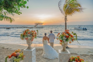 Ceremonia de boda en la playa con una pareja tomándose de las manos, un muelle al fondo y un barco en el agua.