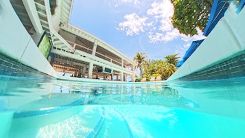 A swimming pool with clear water, a covered area with a ceiling fan, and a view of a building with multiple floors and balconies.