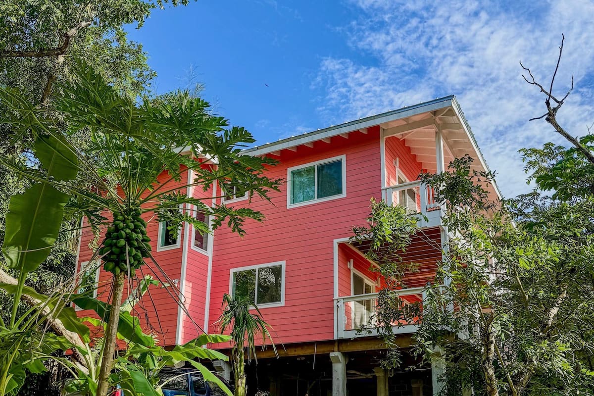 Un edificio rojo de dos pisos con ventanas y balcones enmarcados en blanco, rodeado de abundante vegetación.