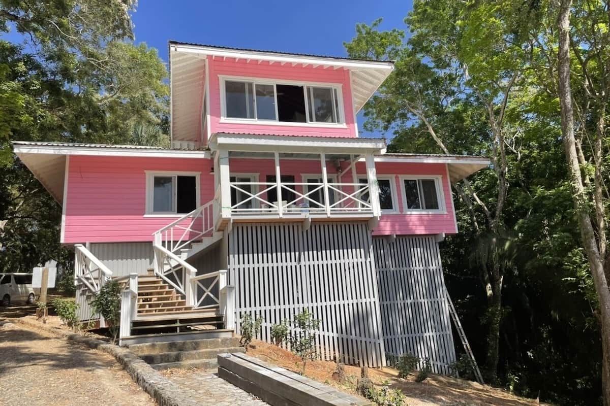 Una casa de dos pisos con fachada rosa, cerca viva blanca y una escalera que lleva a la entrada.