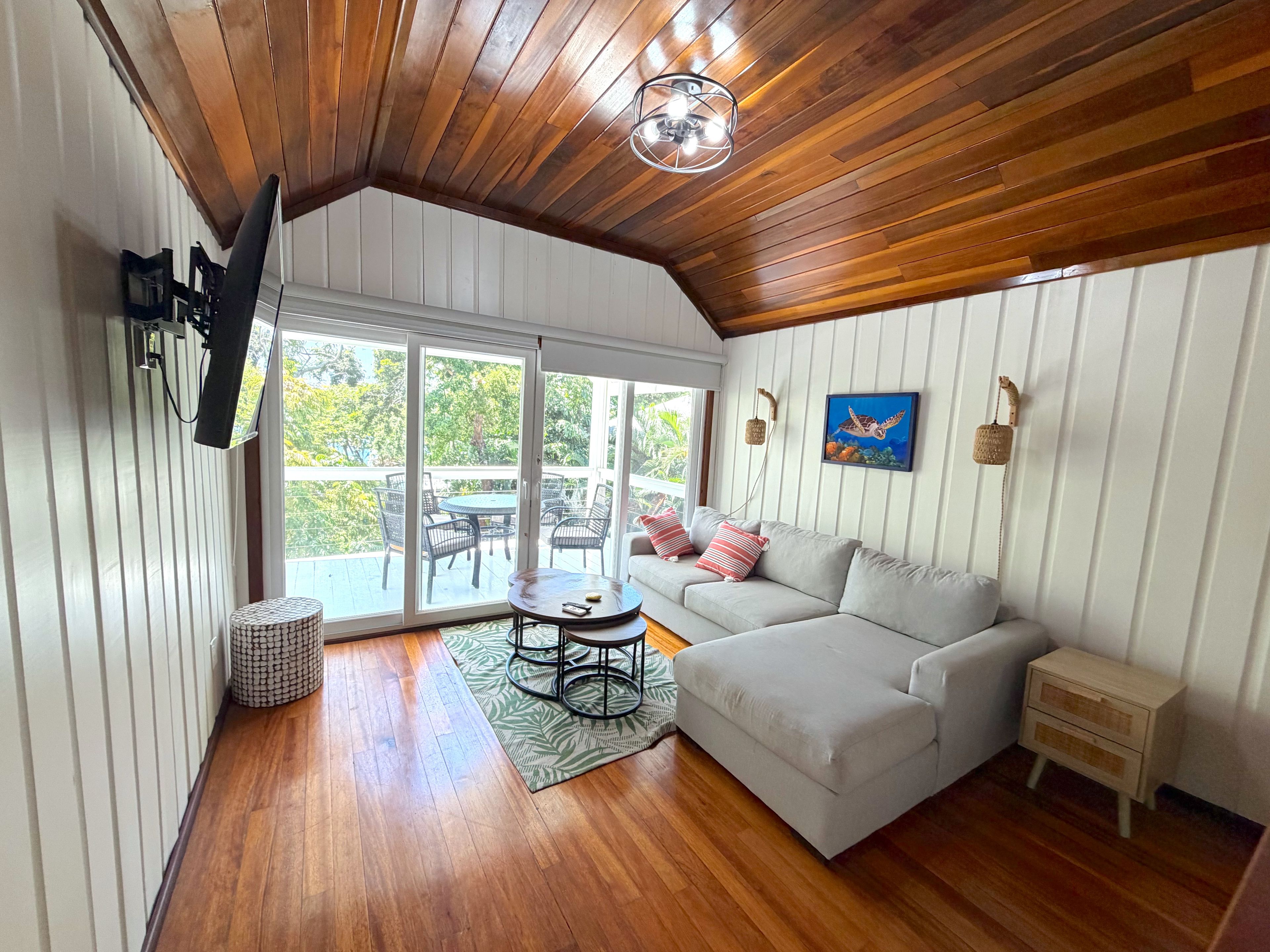 A cozy living room with a wooden ceiling, a sofa, a small table, and a view through large sliding doors to an outdoor patio.