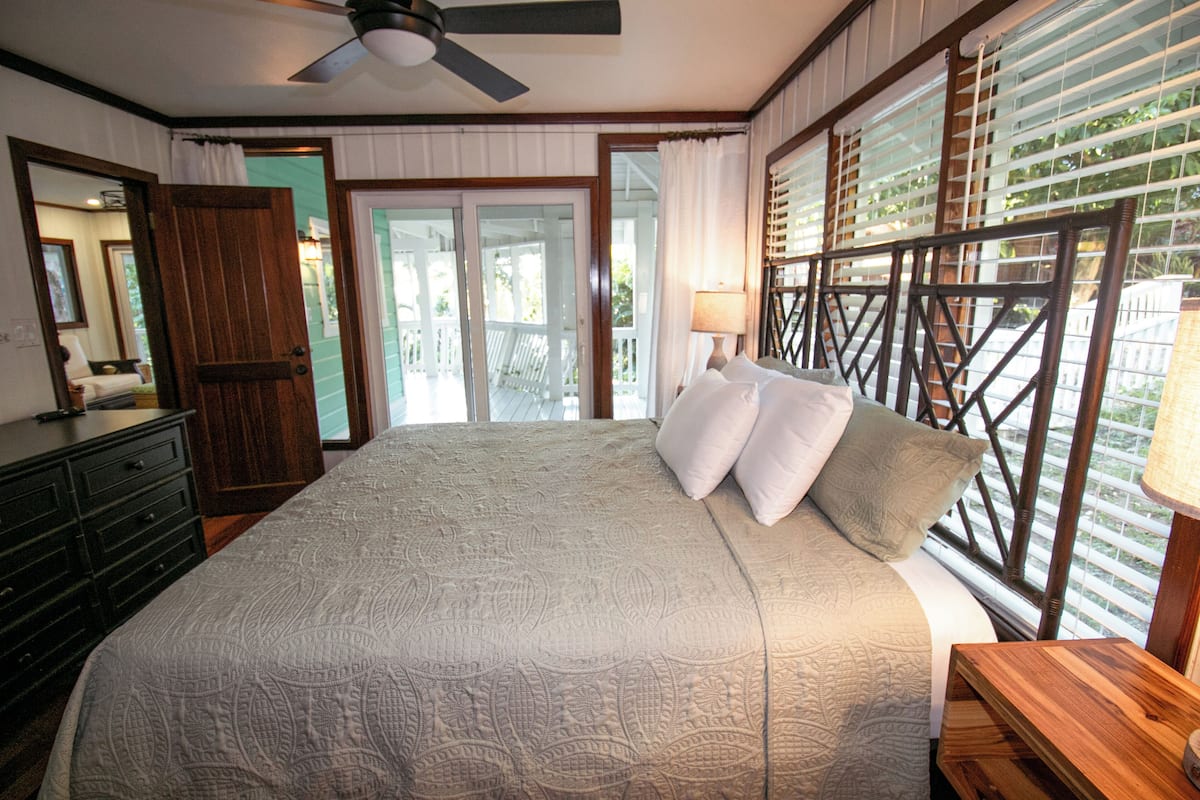 Un dormitorio con una cama grande, un aparador de madera, un ventilador de techo y vistas a un balcón desde la ventana.