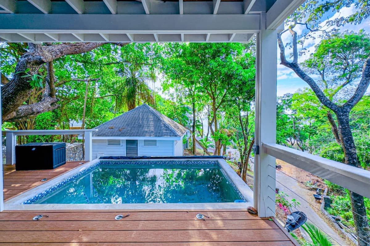 Una piscina techada con un deck de madera, rodeada de exuberante follaje y un pequeño edificio blanco.