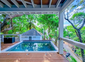 Una piscina techada con un deck de madera, rodeada de exuberante follaje y un pequeño edificio blanco.