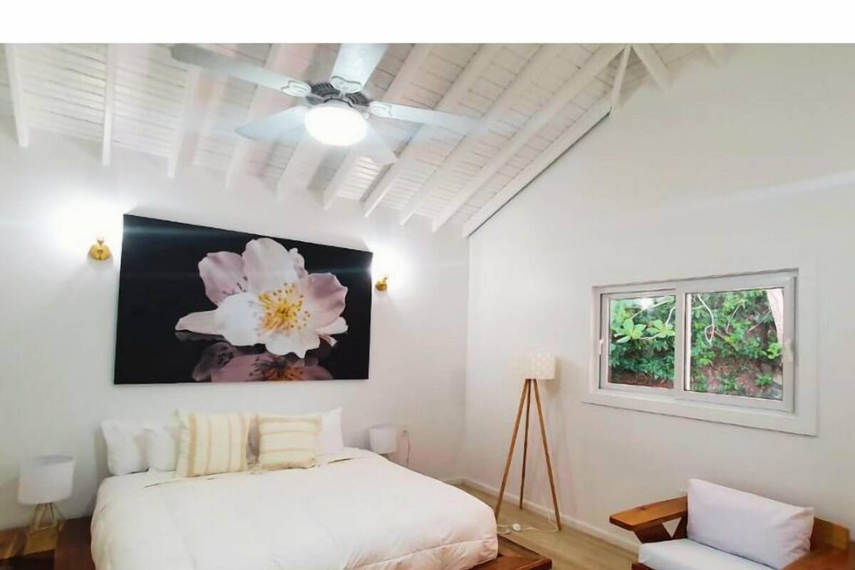 Un dormitorio con cama, ventilador de techo, ventana con vista a vegetación y un cuadro grande de flores en la pared.