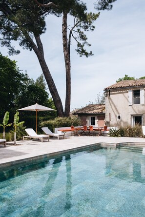 Una piscina con sillas de descanso y una mesa, rodeada de árboles y un edificio con techo de tejas.