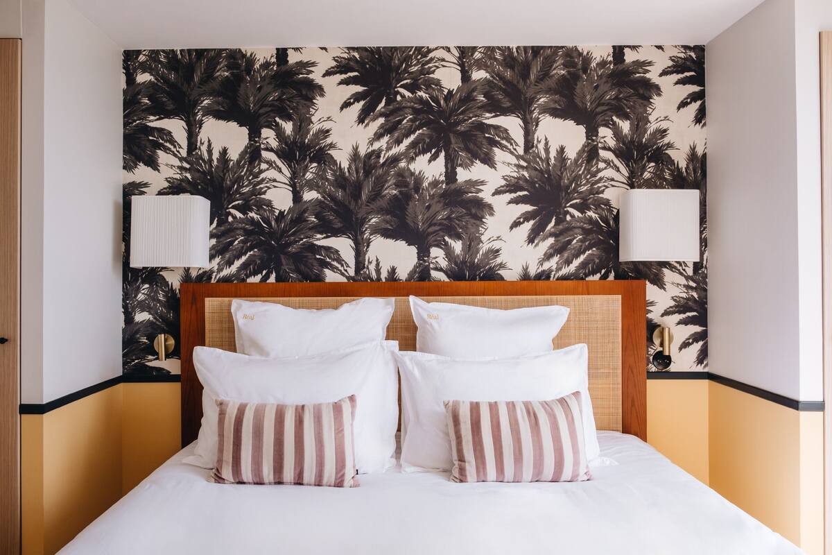 Habitación de hotel con cama, almohadas, cabecero, lámparas fijadas a la pared y una pared con estampado de palmeras.