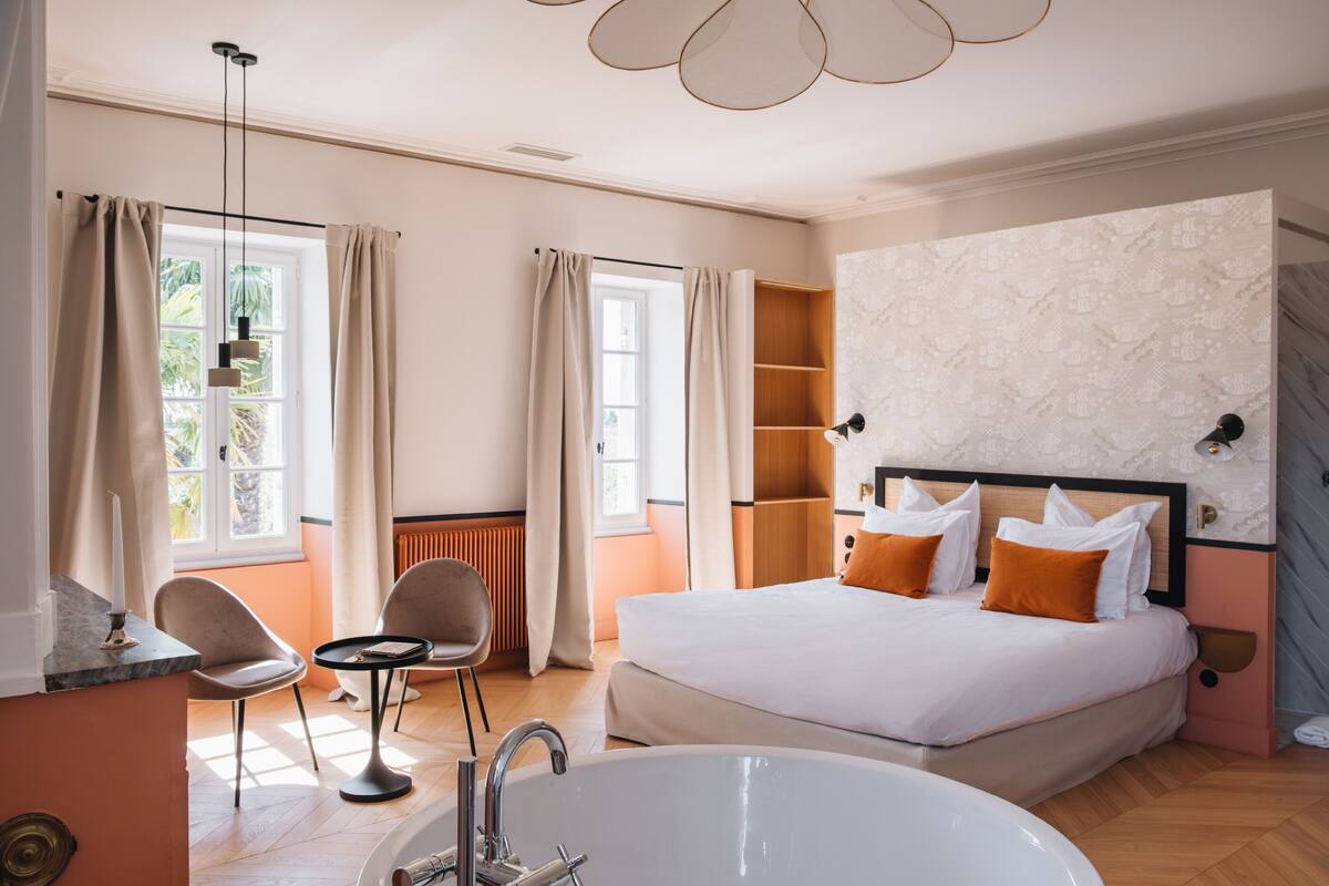 Habitación de hotel con una cama grande, una zona de estar con sillas, una mesita y una bañera, además de un ventanal con cortinas.