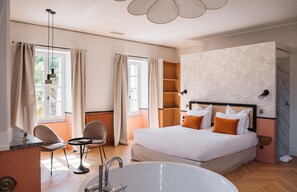 Habitación de hotel con una cama grande, una zona de estar con sillas, una mesita y una bañera, además de un ventanal con cortinas.