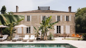 Un espace piscine équipé de chaises longues, d’une table à manger et d’un bâtiment doté d’un toit en tuiles et de volets.