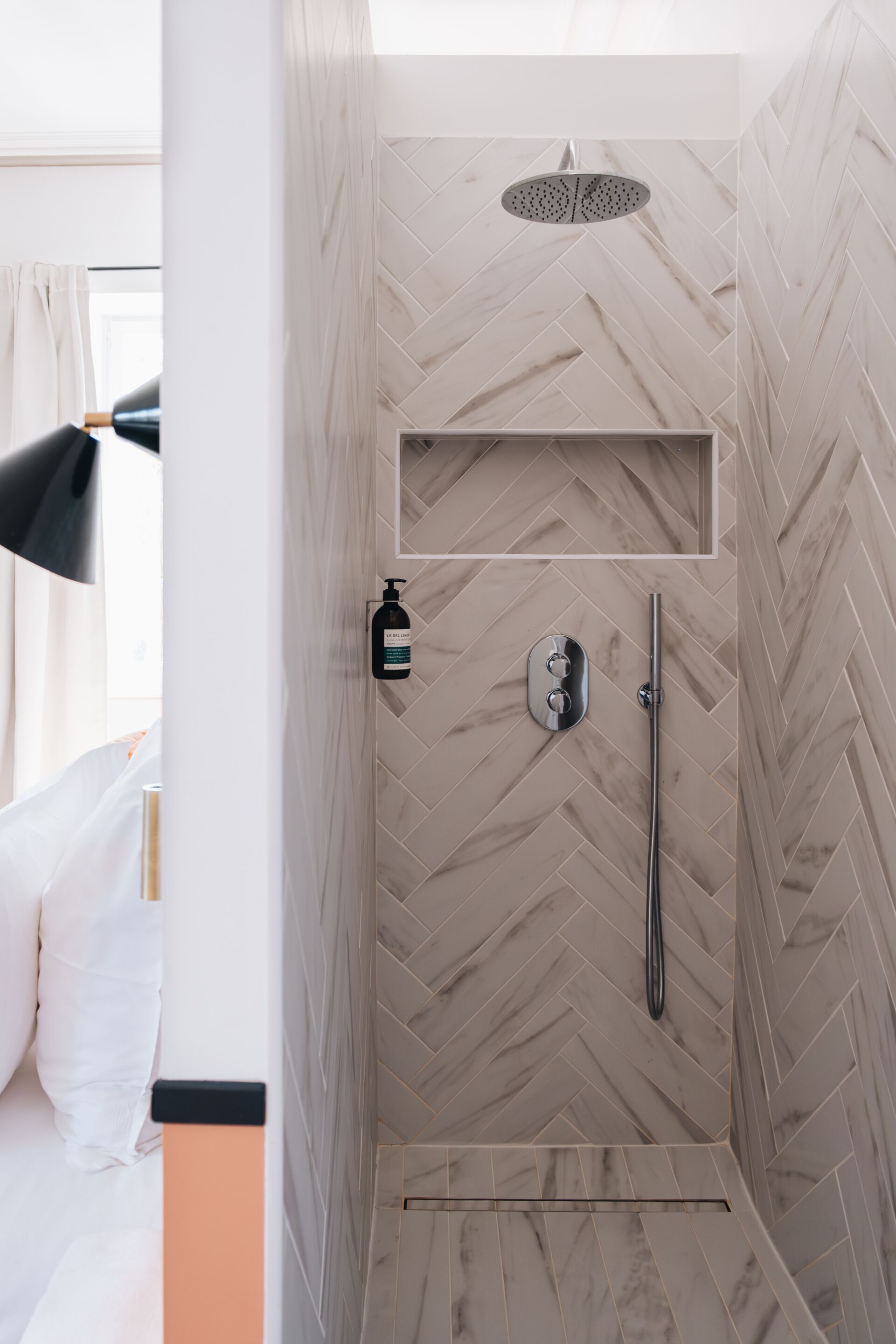 A modern shower with a herringbone tile pattern, a handheld showerhead, and a bottle of shampoo.