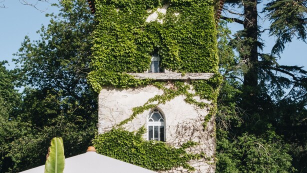 Une tour aux murs recouverts de lierre, avec une porte et une petite fenêtre. On y trouve des fauteuils et un grand parasol devant.