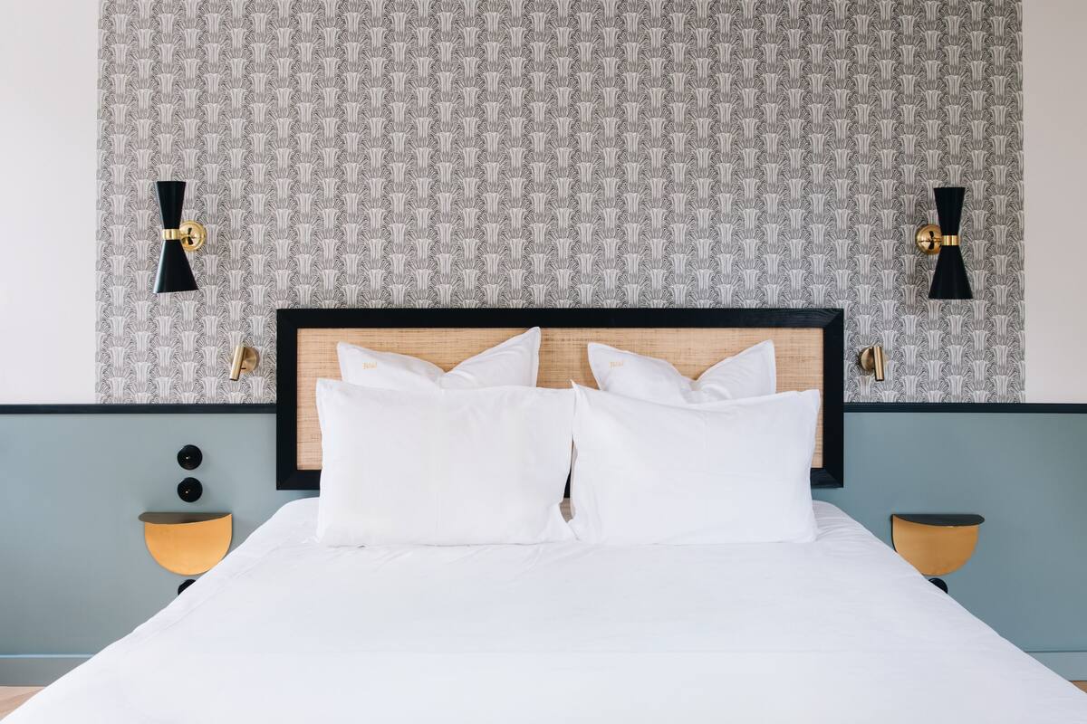 Una cama bien hecha con sábanas blancas y dos almohadas, flanqueada por dos mesitas de noche con lámparas, sobre un fondo de papel tapiz estampado.