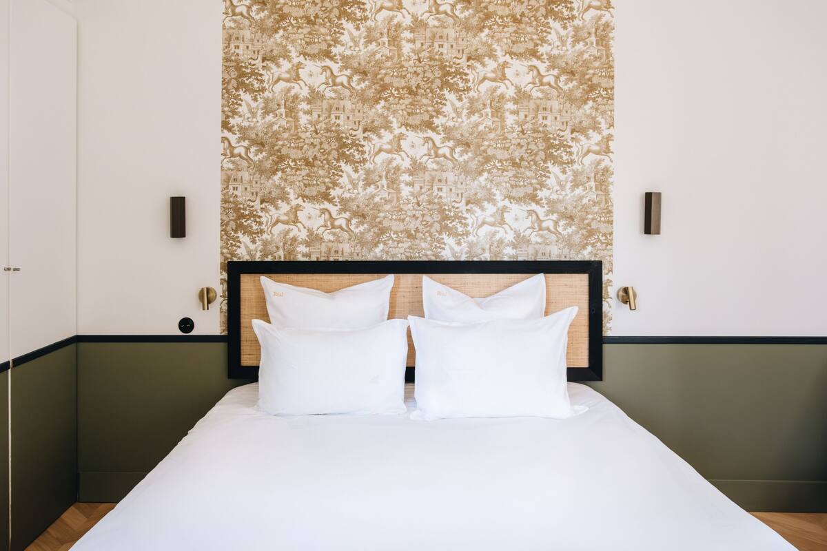 Una cama bien hecha con sábanas blancas y dos almohadas, un cabecero de madera y un papel tapiz estampado con diseño floral.
