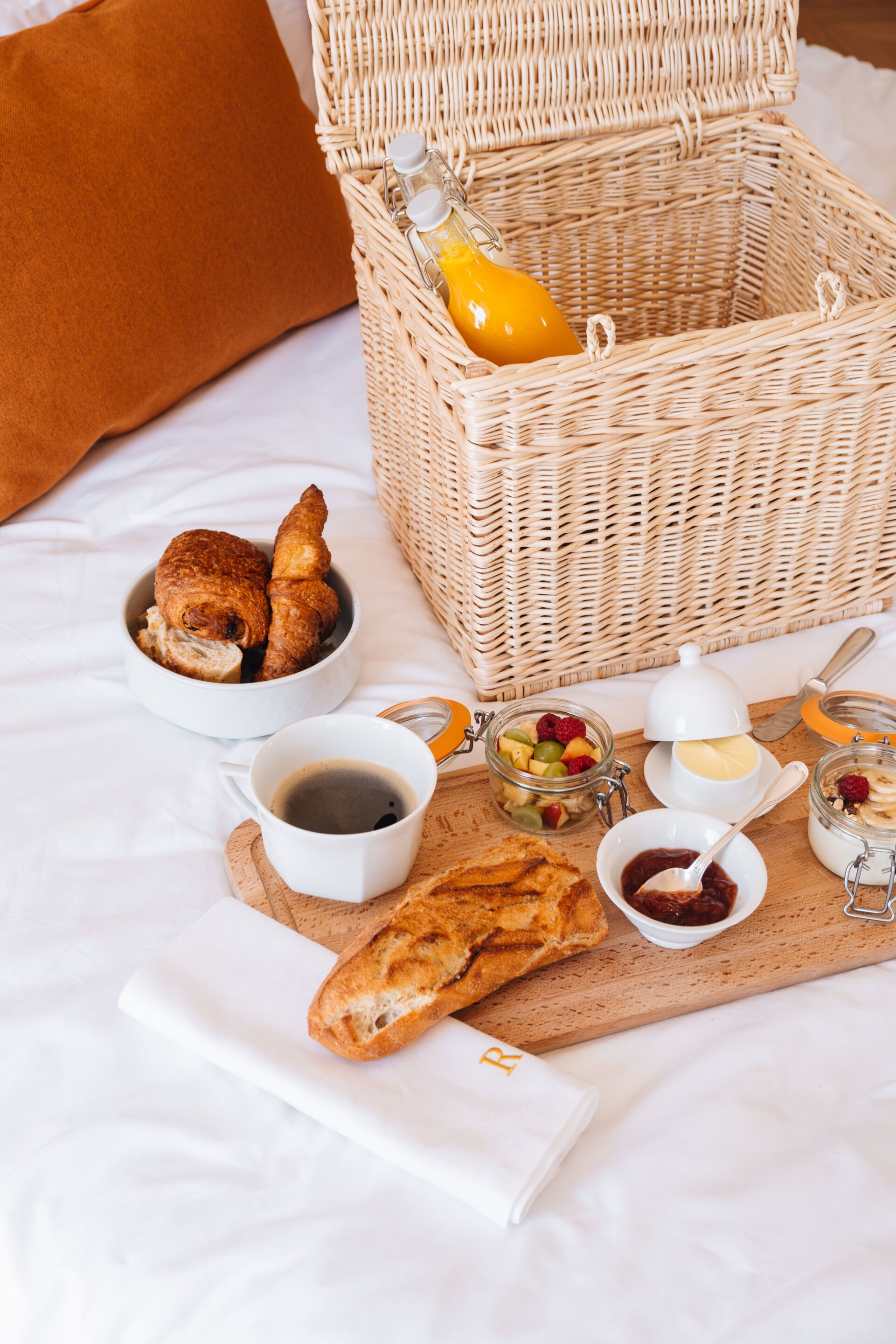 A breakfast setup with bread, pastries, a basket, and various condiments on a bed.