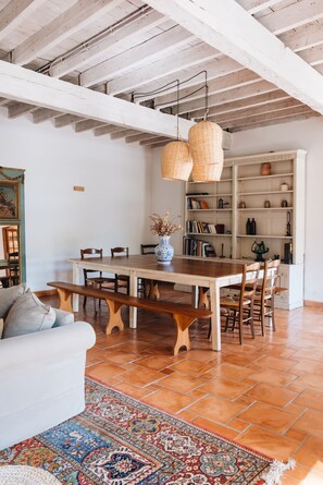Un comedor amplio con una mesa y sillas de madera, un banco, un sofá blanco, una alfombra estampada y una estantería llena de libros y objetos decorativos.