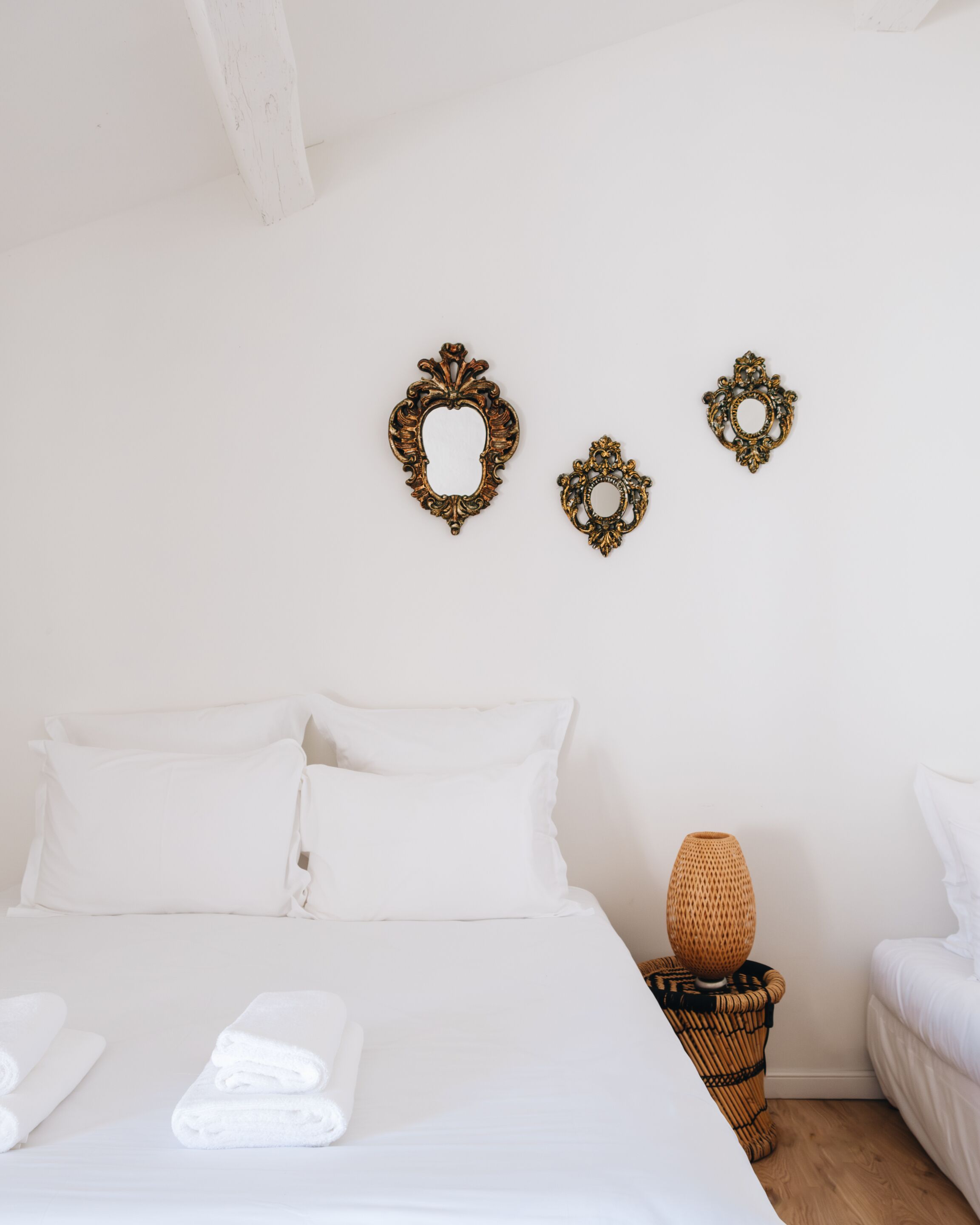 Een net opgemaakt bed met witte lakens en kussens, een klein geweven mandje met een vaas, en twee sierlijke wanddecoraties.