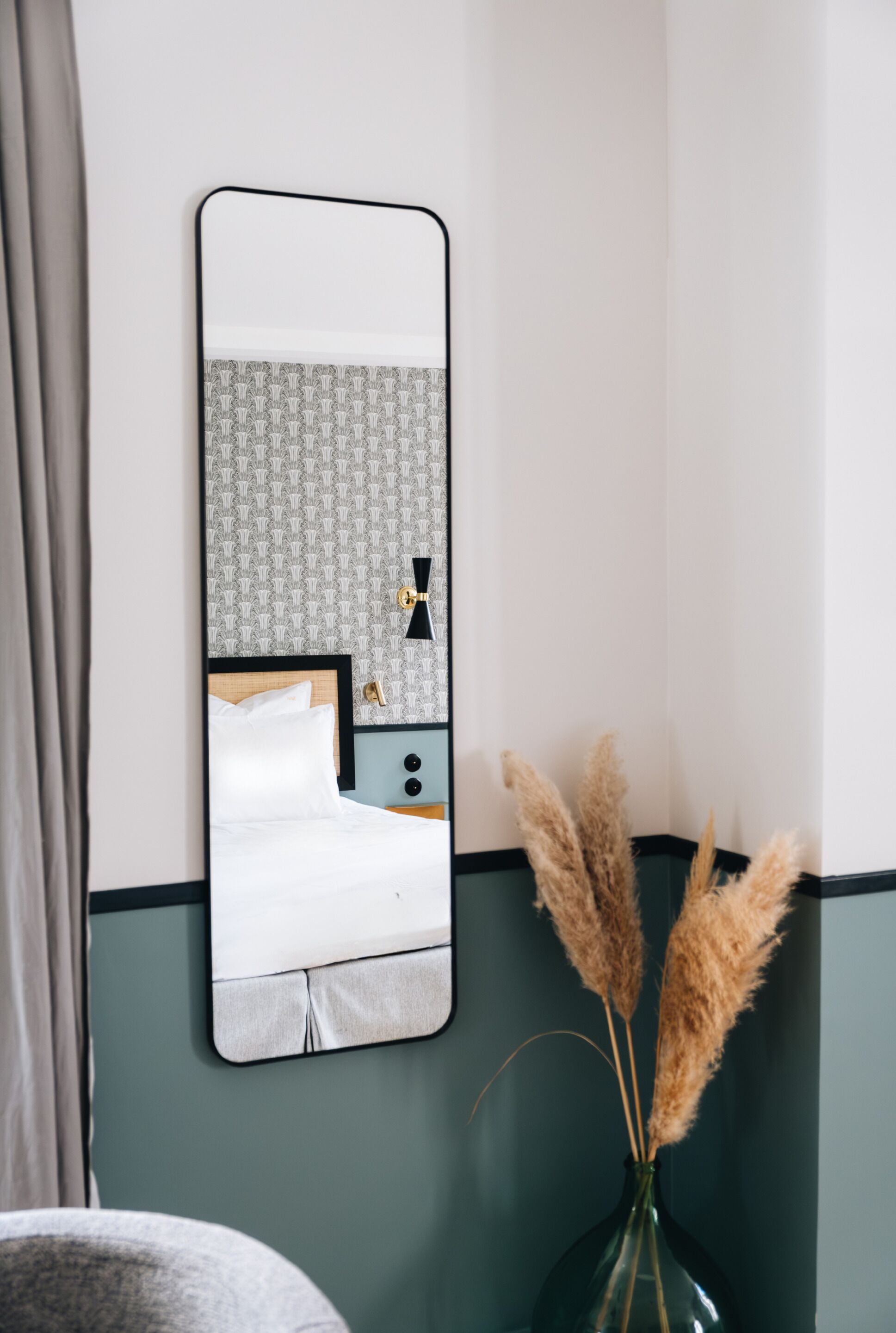 A mirror reflecting a bed, a nightstand, and a vase with pampas grass.