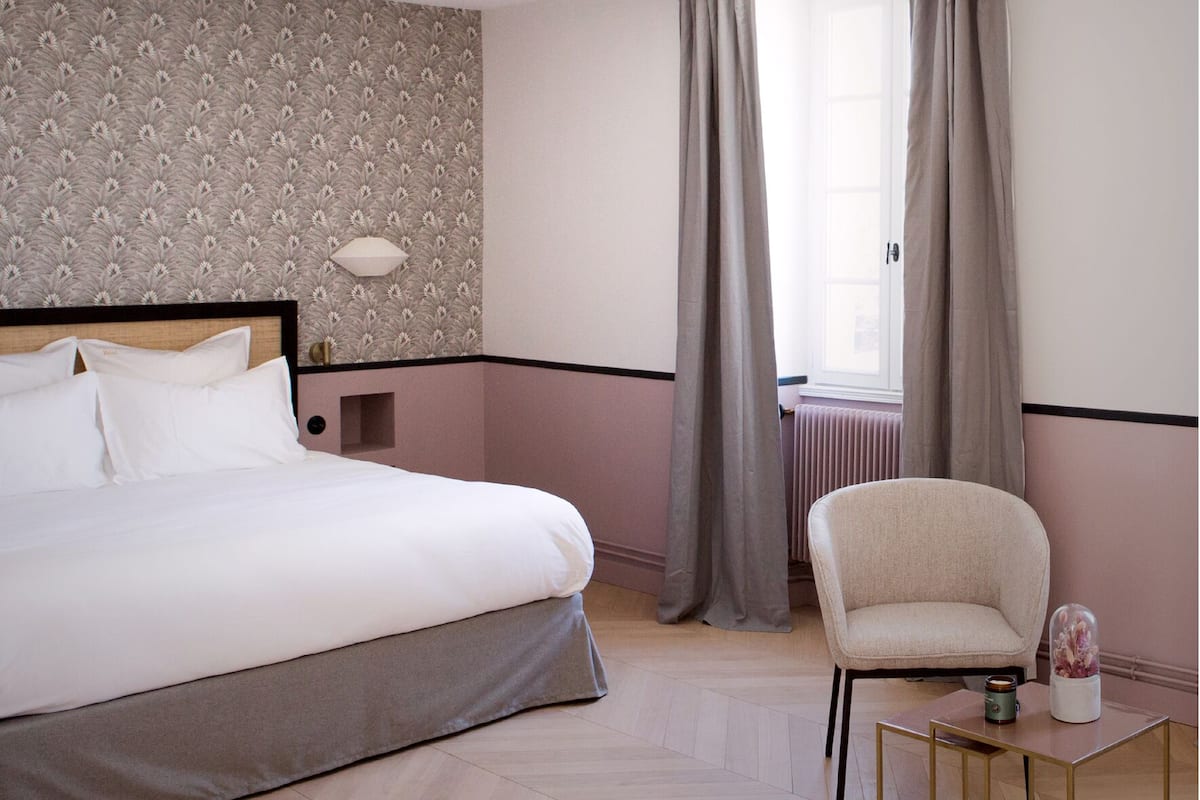 Una habitación de hotel con una cama, una silla, una mesita con lámpara y una ventana con cortinas.