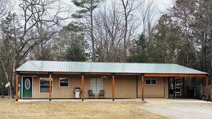 A single-story building with a green metal roof, a front porch with two chairs, and a small attached garage.