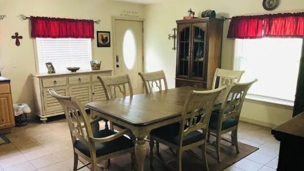 A dining room with a wooden table and six chairs, a cabinet, and a window with red curtains.
