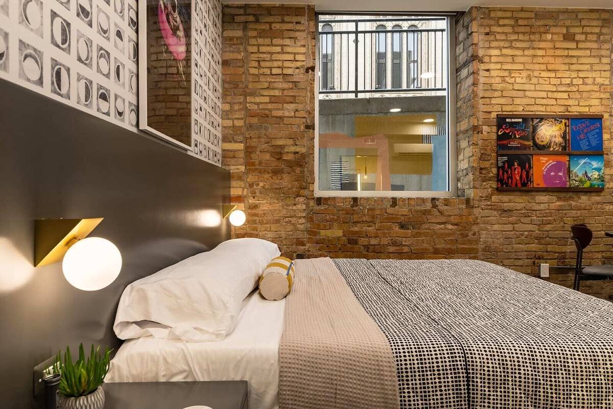 Un dormitorio moderno con pared de ladrillo, un ventanal grande, una cama con ropa blanca y obras de arte colgadas en la pared.