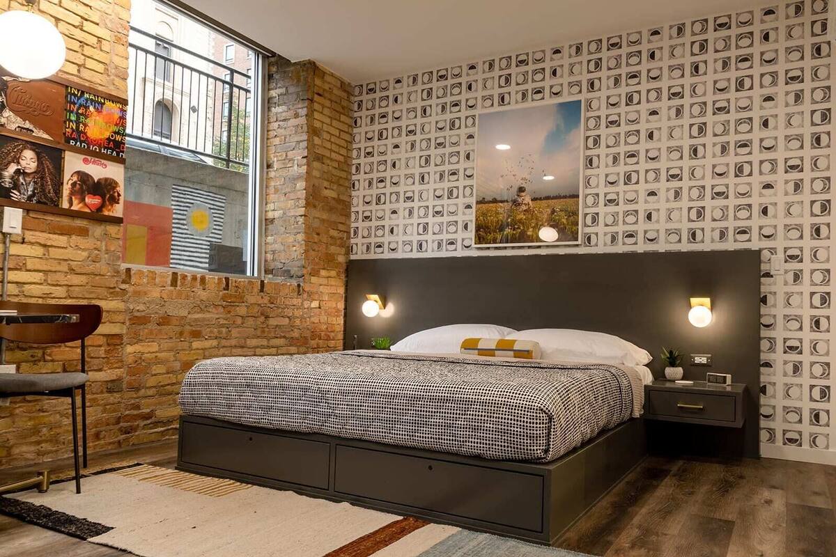 Un dormitorio con pared de ladrillo, una cama con ropa de cama estampada, una mesita de noche y luces fijadas a la pared.