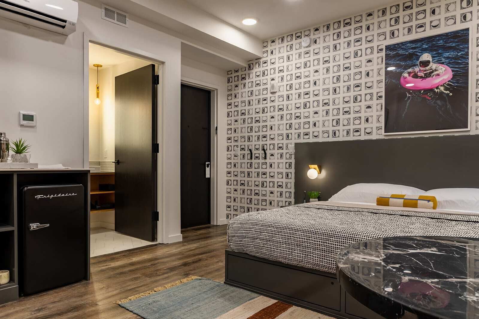 A modern hotel room with a bed, a desk with a refrigerator, a wall with circular patterns, and a poster of an astronaut.