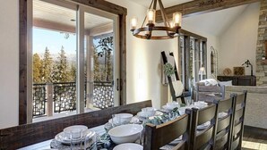 A dining area with a wooden table set for a meal, large windows with a view of trees, and a rustic chandelier.