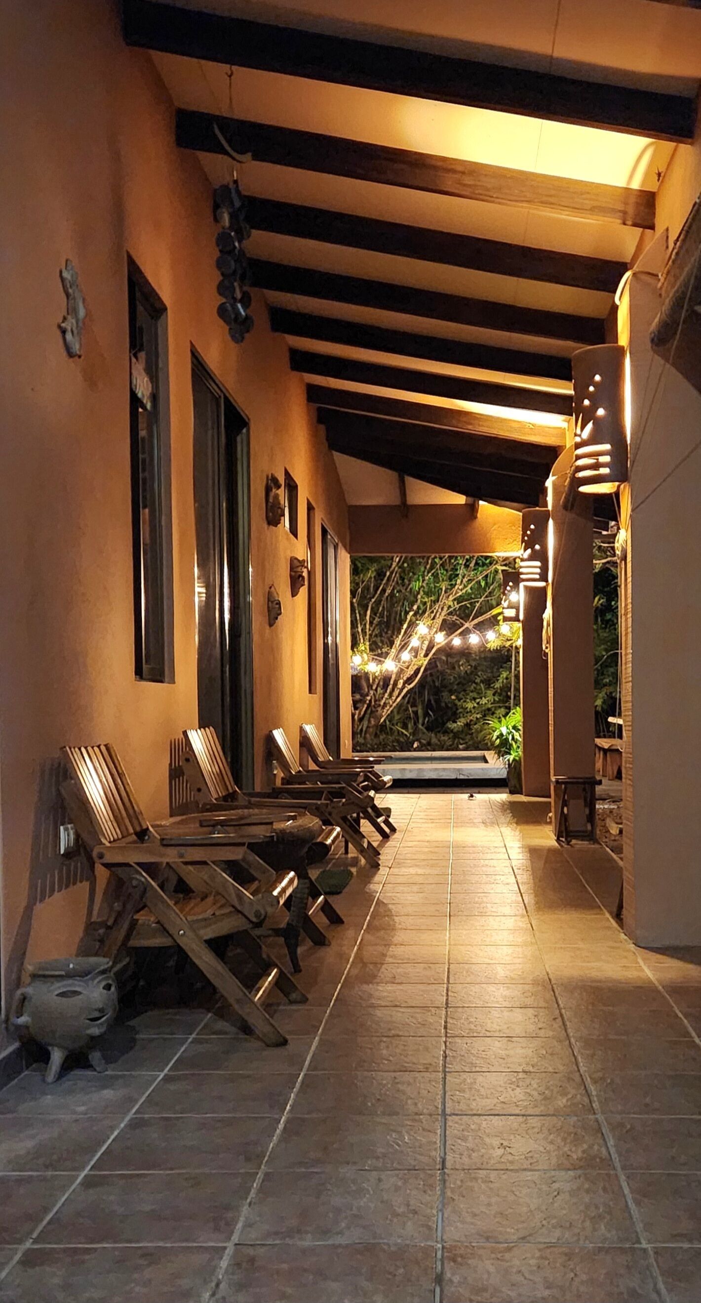 A covered outdoor seating area with wooden chairs and tables, a tiled floor, and string lights.