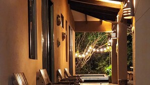 Een overdekt terras met houten stoelen en tafels, een betegelde vloer en lichtslingers.