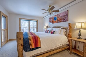 A bedroom with a wooden bed frame, a ceiling fan, a large window, and a painting on the wall.