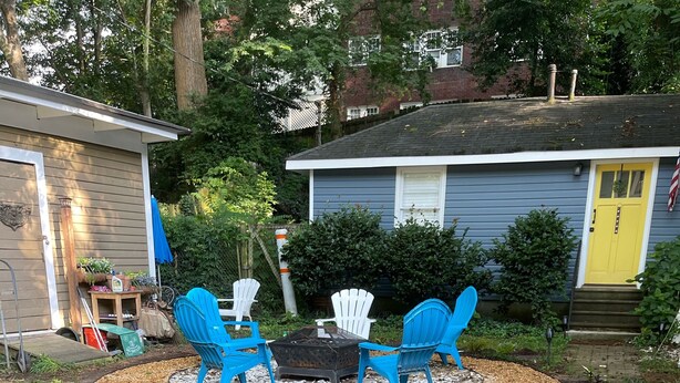 Un jardin avec un foyer et des chaises bleues. On aperçoit des plantes et un abri de jardin au fond.