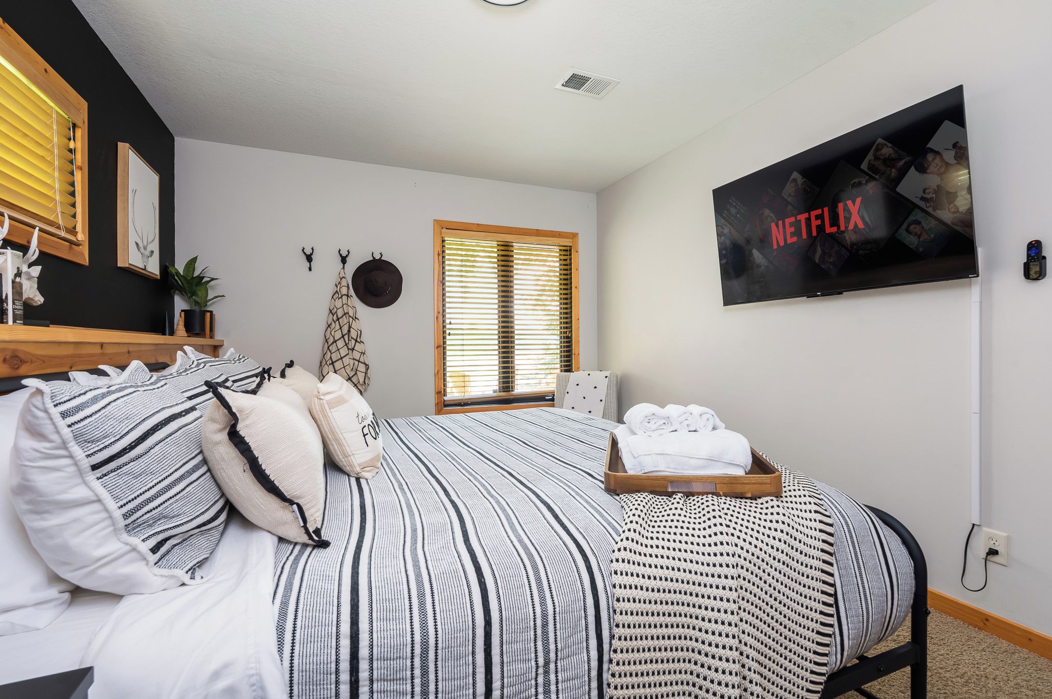 Un dormitorio con cama, almohadas, una bandeja con toallas, un televisor empotrado mostrando el logotipo de Netflix, una ventana con persianas y una pequeña planta.