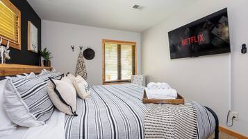 Un dormitorio con cama, almohadas, una bandeja con toallas, un televisor empotrado mostrando el logotipo de Netflix, una ventana con persianas y una pequeña planta.