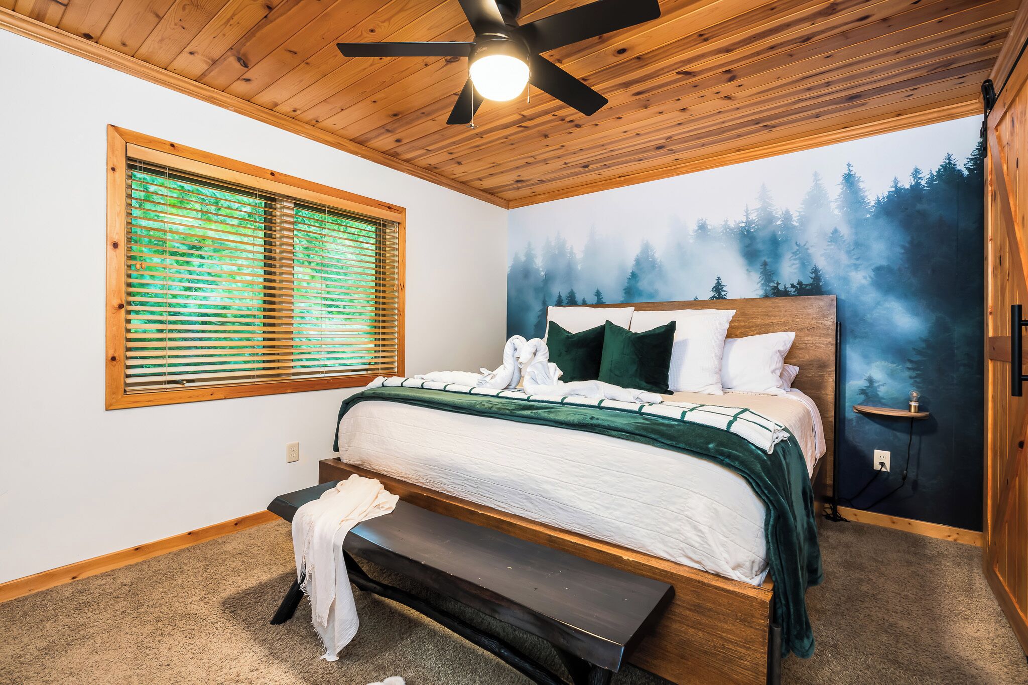 A bedroom with a bed, bench, ceiling fan, window with blinds, and a mural of a forest.