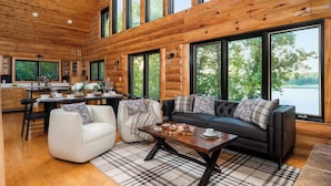 A spacious living area with a large sofa, a coffee table, and a dining table. There are multiple windows providing views of the outdoors. - Summer oasis w/NEW HOT TUB overlooking Fawn Lake with private beach, SUPs/Kayaks (Crosslake)