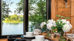 A dining table set with white dishes and glasses, a vase with white flowers, and a view of a lake through large windows. - Summer oasis w/NEW HOT TUB overlooking Fawn Lake with private beach, SUPs/Kayaks (Crosslake)