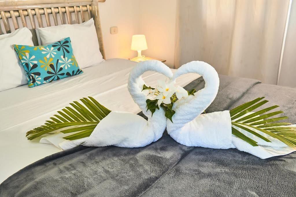 A neatly made bed with white and blue pillows, a heart-shaped towel decoration, and a bedside lamp.