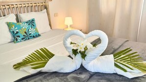 A neatly made bed with white and blue pillows, a heart-shaped towel decoration, and a bedside lamp.