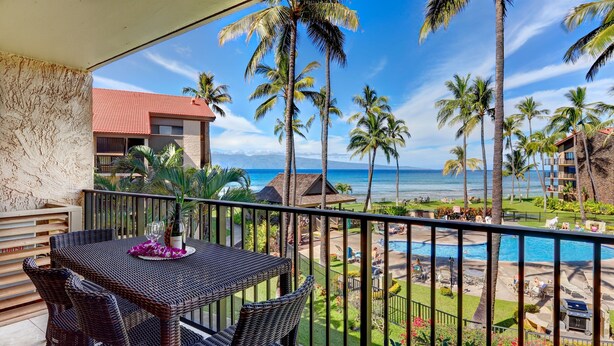 A balcony with wicker furniture, a table with a bottle and glasses, and a view of a pool and beach with palm trees. - Stunning Ocean Views From Highly Upgraded 2 Bedroom Condo: Papakea K303 (Lahaina)