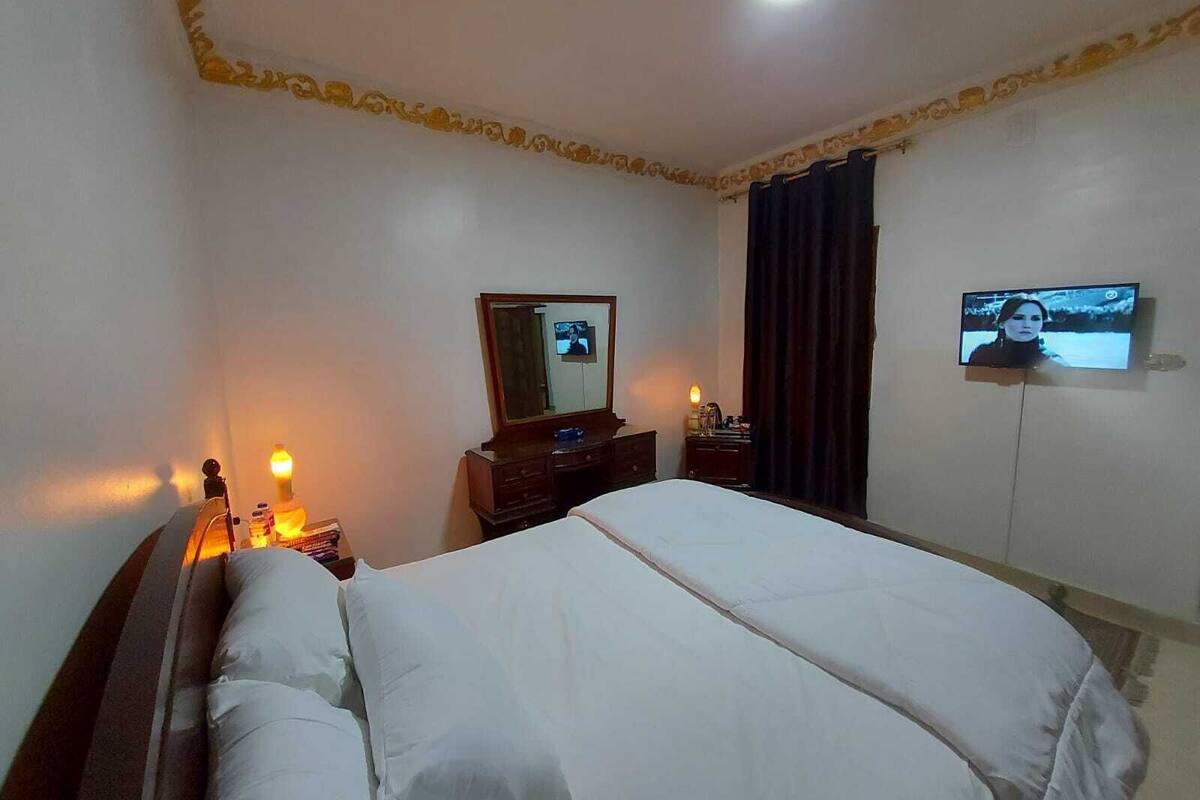 A hotel room with a bed, a dresser, a mirror, a television, and a nightstand with a lamp.