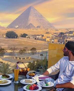 Una mesa de comedor con diversos platos y un vaso de jugo de naranja, con una pirámide y un paisaje urbano de fondo.