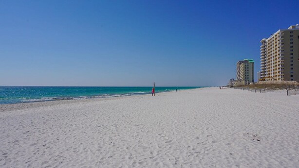 A wide sandy beach with a clear blue sky, a few people, and high-rise buildings in the background.