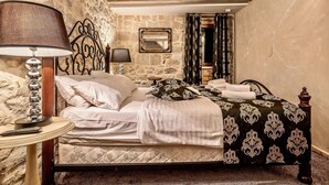Une chambre avec un mur en pierre, un lit recouvert d’une couvre-lit à motifs, une table de chevet avec une lampe et un miroir accroché au mur.