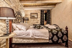 Un dormitorio con pared de piedra, una cama con colcha estampada, una mesita de noche con lámpara y un espejo en la pared.