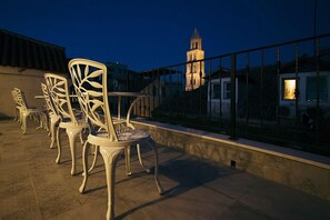 Mobiliario exterior con sillas y una mesa, barandilla y un edificio con torre al fondo.